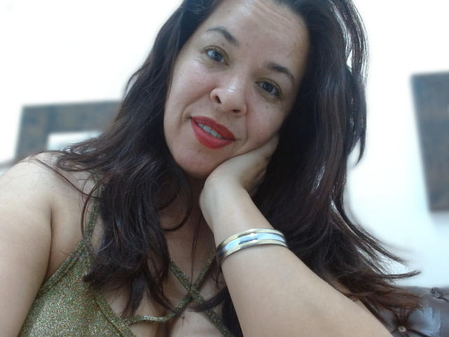 LizethBells with dark brown hair, red lipstick, green eyes, and a gold sparkly top, resting her head on her hand, wearing a silver bracelet, smiling.