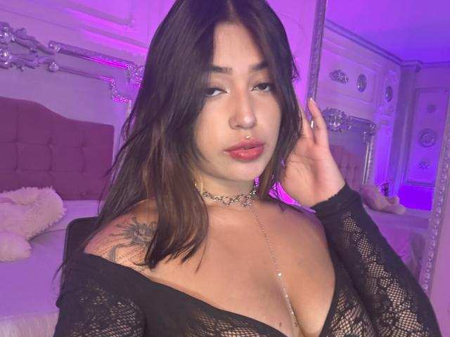 KatianPierce with dark hair, red lipstick, black lace top, choker, tattoo on shoulder, hand in hair, looking at camera, pink-purple room, visible cleavage, small nose ring.