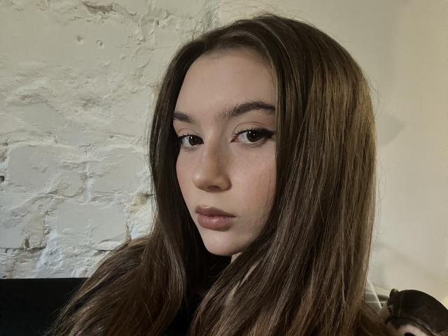 BarbaraCrazy has long brown hair, dark eyeliner, and a neutral expression, with pale skin and subtle makeup. Her tits aren't visible. She's looking slightly to the side.