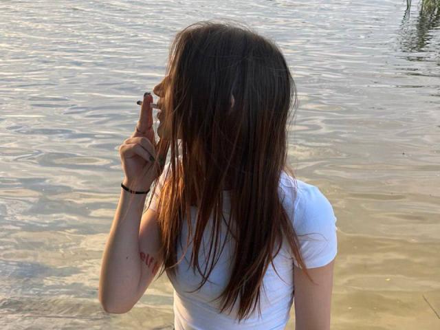 BarbaraCrazy with long brown hair, white shirt, standing in shallow water, holding cigarette to mouth, slightly bent over, wearing black bracelet, water reflecting light.