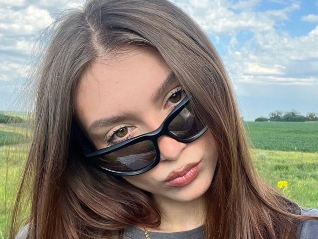 BarbaraCrazy with brown eyes, wearing black sunglasses, has long brown hair and pouty pink lips, looking slightly down, in a green field under a blue sky with clouds.