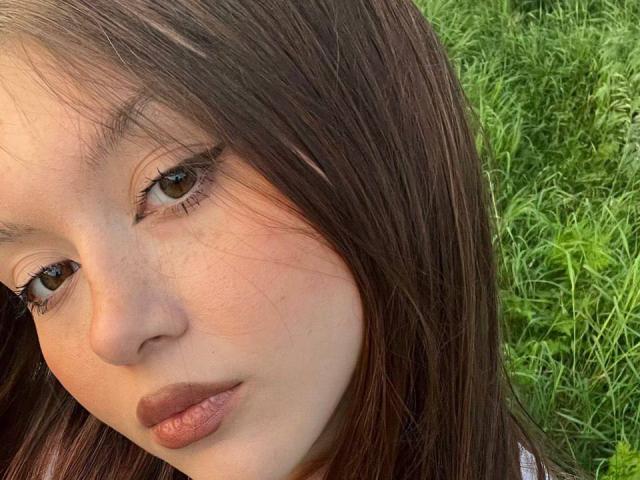 BarbaraCrazy's face, brown eyes, long brown hair, light skin, brown lipstick, slight freckles, looking to the side, green grass in the background, soft natural light.