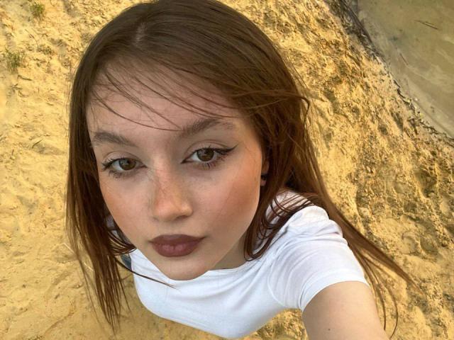 BarbaraCrazy, young woman with brown eyes, freckles, and brown lipstick, wearing a white shirt, taking a selfie from above, showing her face and upper chest.
