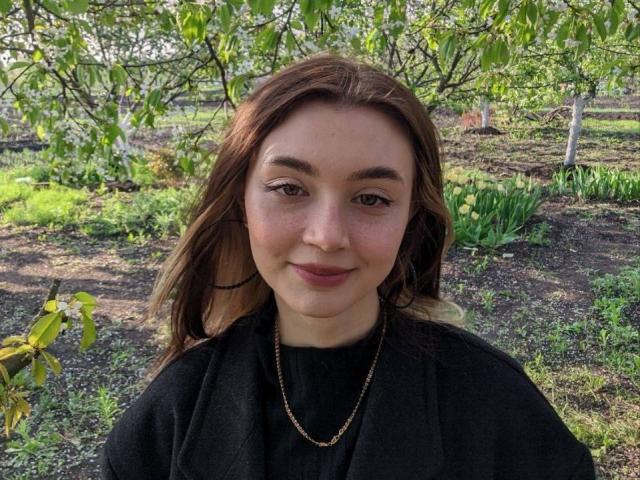 BarbaraCrazy with brown hair, fair skin, and a subtle smile, wearing a black shirt and gold necklace, has small tits and a cute face, standing outdoors.