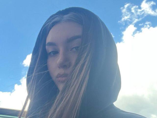 BarbaraCrazy with long brown hair in a black hood, blue sky, and white clouds, looking down with a slightly pouty expression. Soft sunlight highlights her face.