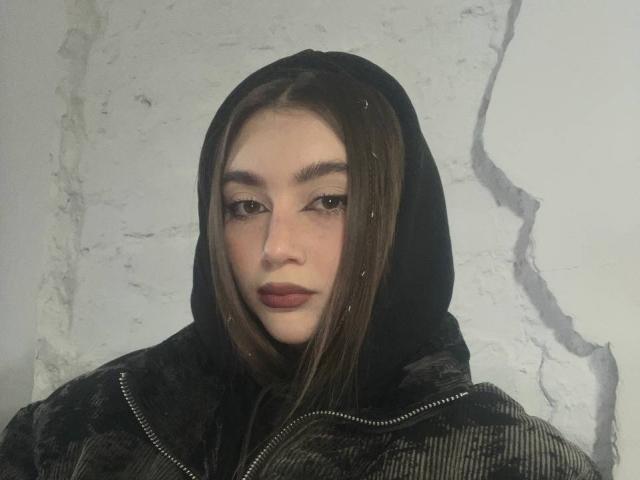 BarbaraCrazy with straight brown hair, black hood, and dark red lipstick, wearing a black puffy jacket, has a serious, slightly sad expression, with small white specks on her face.