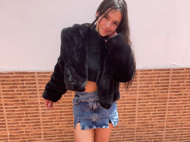 MarietteJones with long brown hair, black fur jacket, black crop top, and blue denim skirt, leaning against a brick wall, smiling, hand near face, small ass, cute.