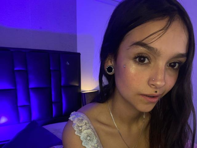 MarietteJones has long brown hair, brown eyes, and multiple facial piercings including a nose ring and lip ring. She's wearing a lace off-shoulder top, with blue bedroom lighting.