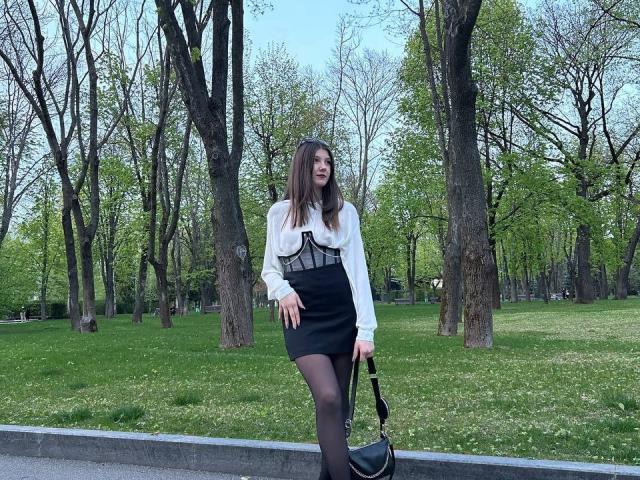 LissaNight has long brown hair, a white blouse with black mesh, a black skirt, black stockings, and holds a black bag. She's standing outdoors, smiling.