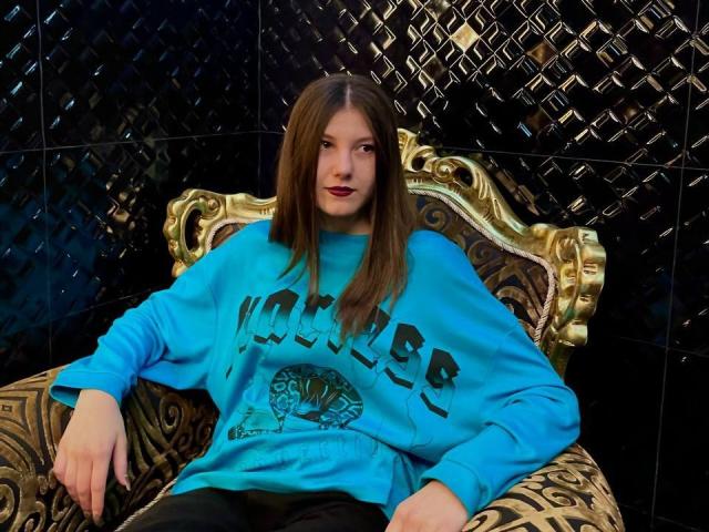 LissaNight with long brown hair, blue "Voomer" sweatshirt, and black pants, sitting on an ornate gold chair, relaxed, with a slight smirk, in a dark room.