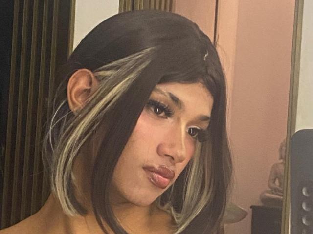 HillaryRosee tem cabelo castanho escuro com mechas loiras, sobrancelhas grossas, cílios longos e batom rosa. Ela está olhando para baixo com uma expressão levemente emburrada em uma selfie no espelho..