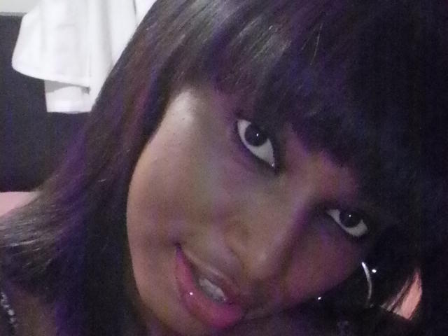 AnaSexy has dark purple hair, black eyeliner, and a slight smile with glossy pink lips, showing her teeth. Her eyes are looking directly at the camera.