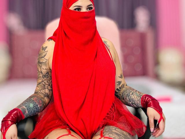 MalakCote sits in a chair, wearing a red mask, red dress, and red gloves, exposing tattooed arms and red-tinted thighs, with a red headscarf covering her face.