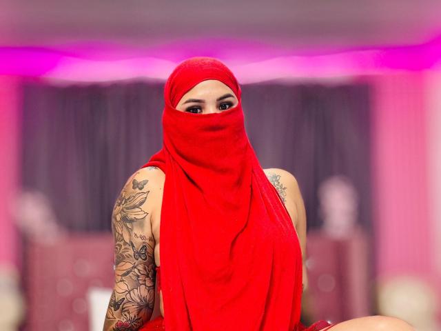 MalakCote wearing a red headscarf and matching mask, showcasing tattooed arms and looking at the camera, with pink and gray background. Boobs partially covered, pussy not visible.