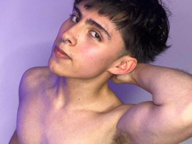 PepperTwink, shirtless, with short dark hair, a nose ring, and a slight smirk, has a muscular arm raised, showing a hairy chest, and a relaxed, confident expression.