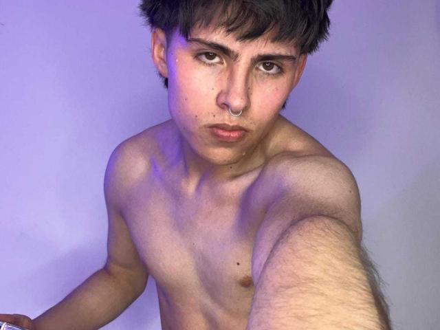 PepperTwink has a serious face, dark short hair, a nose ring, and is shirtless with a slight purple light on his hairy chest and arm. His hand is reaching toward the camera.