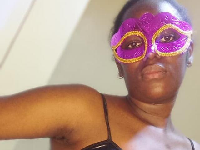QuinnLily tem a pele marrom escura e usa uma máscara de carnaval roxa e dourada, mostrando uma expressão séria com um leve franzir de lábios. Ela veste uma blusa preta com alças finas..