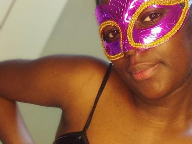QuinnLily has a shiny purple mask with yellow glitter, brown eyes, and a dark brown skin tone. She's wearing a black strap top, and smiling slightly.