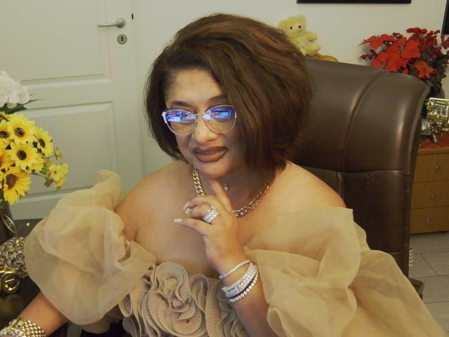 Elegante pero provocativa, con cabello castaño corto, gafas y un vestido marrón de corte asimétrico que deja ver sus senos, con un anillo en el dedo, llevando joyas, sentada en una silla marrón..