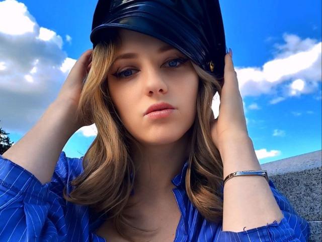 JoseFairy with blue eyes, wearing a shiny black cap and a blue pinstriped shirt, hands in cap, looking serious, brown wavy hair, wrist bracelet, bright blue sky.