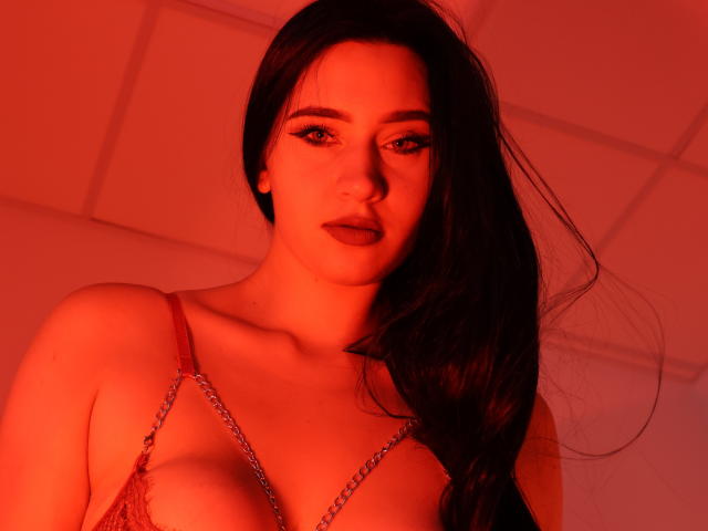 MaryLori with red-lit skin, long black hair, and a low-cut, chain-patterned top, showing her tits and a serious, seductive expression.