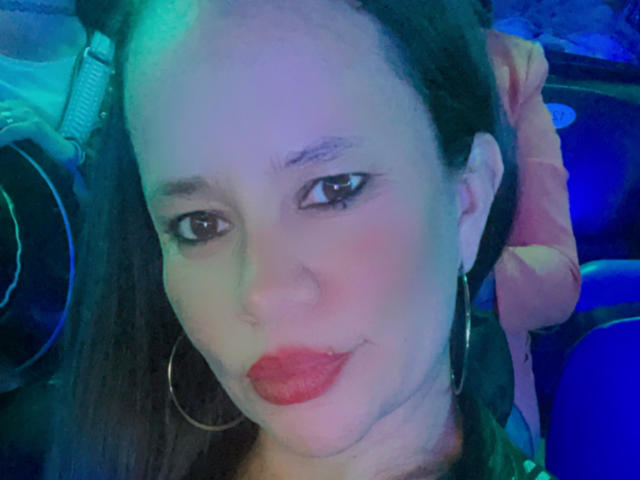 CharllotI a les lèvres rouges, les yeux marron et les cheveux noirs tressés avec un ruban rose. Elle porte des boucles d'oreilles cerceaux et une lumière verte-bleue éclaire son visage..