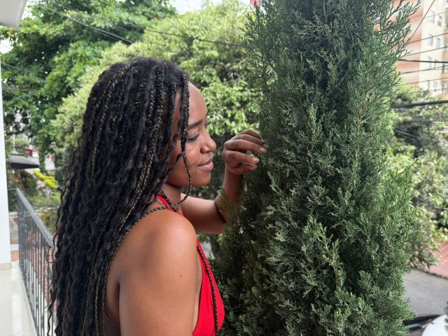 NaomiReyes with long black braids, red top, touching a tall green tree, eyes closed, calm expression, outdoor balcony, lush greenery in background, soft natural light, relaxed.
