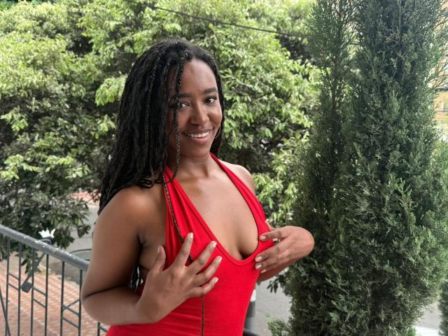 NaomiReyes with dark skin, smiling, wearing a red halter top, hands on her tits, standing outdoors, greenery in background, long black braided hair, cute, confident.