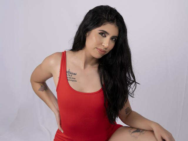 MiluSky with long black hair, wearing a red tank top, has tattoos on her left arm and chest, sitting with hands on knees, smiling slightly, showing small tits.
