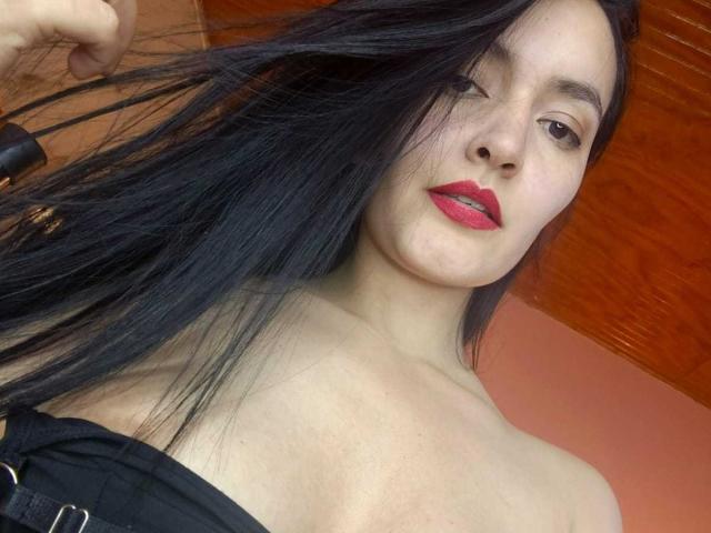 AriaBonnet has long black hair, red lipstick, and is holding her hair with a hand, showing bare shoulders and a black strap. Her tits are partially visible.