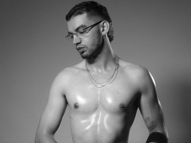 MichaelMagno's shirtless, sweaty, muscular torso with shiny skin, small nipples, tattoo on right arm, chain necklace, glasses, and short curly hair, looking down with a serious expression.