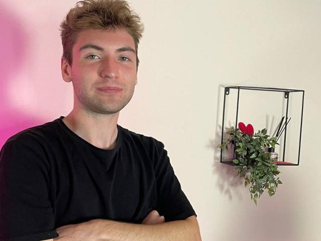 JosephBest, with crossed arms, wearing a black t-shirt, has short, messy brown hair and a slight smile. A wall shelf with a red heart, plant, and reeds is behind.