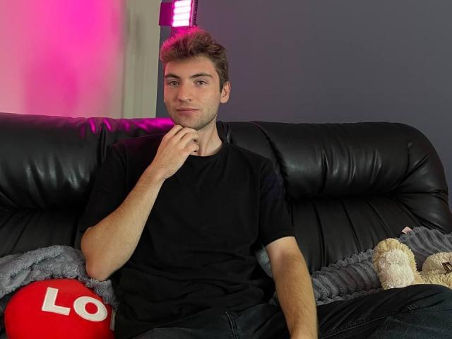 JosephBest, with short brown hair and a slight smile, sits on a black leather couch, wearing a black shirt and dark jeans, with a hand on his chin.