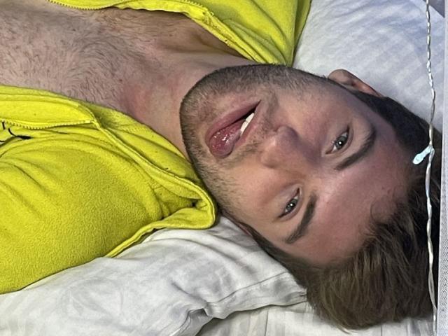 JosephBest is lying on a pillow, wearing a yellow zip-up, his tongue slightly out, looking up with blue eyes and a slight smile, with a white earbud.