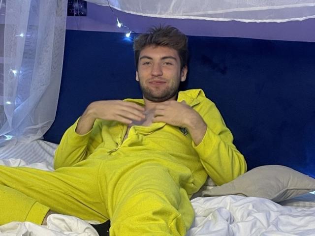 JosephBest with a light beard, wearing a yellow zip-up pajama suit, sitting on a bed, hands near chest, smiling, light brown hair, white bed sheets, blue wall.