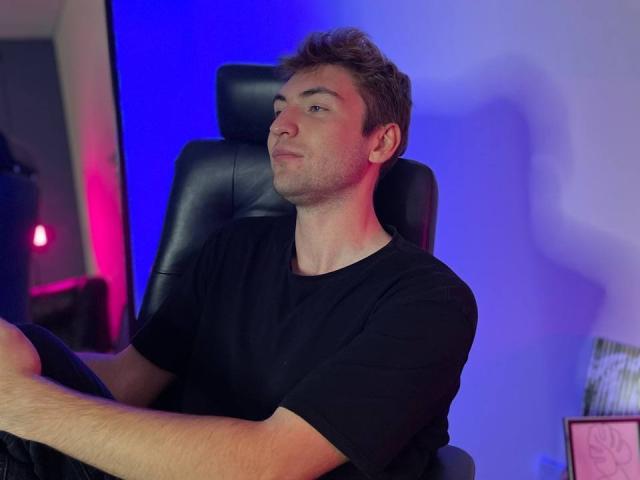 JosephBest is a cute dude with short brown hair, wearing a black t-shirt, sitting in a black leather chair, looking to the side with a relaxed expression.