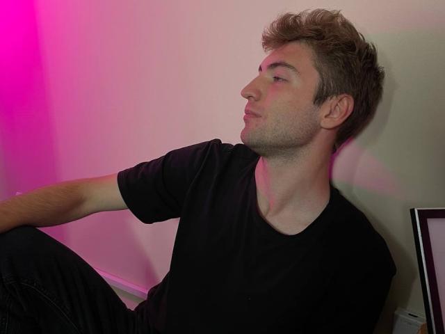 JosephBest is a cute, blonde dude with a slightly messy haircut, wearing a black t-shirt and black pants, sitting with a relaxed, side-profile gaze, under pink lighting.