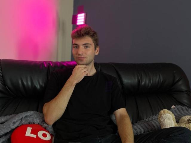 JosephBest with short brown hair, black t-shirt, and blue jeans, sitting on a black leather couch, hand on chin, pink neon light in background, gray blanket and stuffed toy.