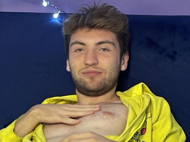 JosephBest with messy brown hair and beard, yellow hoodie open, exposing chest with small nipple, hand resting on it, slightly smiling, string lights in background.