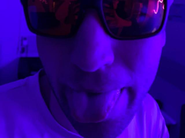 JosephBest's face, illuminated in purple, with tongue out, wearing reflective sunglasses, white shirt, and a black necklace, in a slightly blurred, low-light image.