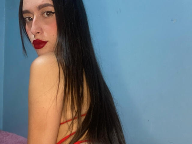 SarahLuxe with long black hair, red lipstick, and red strappy top, looking over her shoulder, showing her bare back and small ass. Blue wall in background.