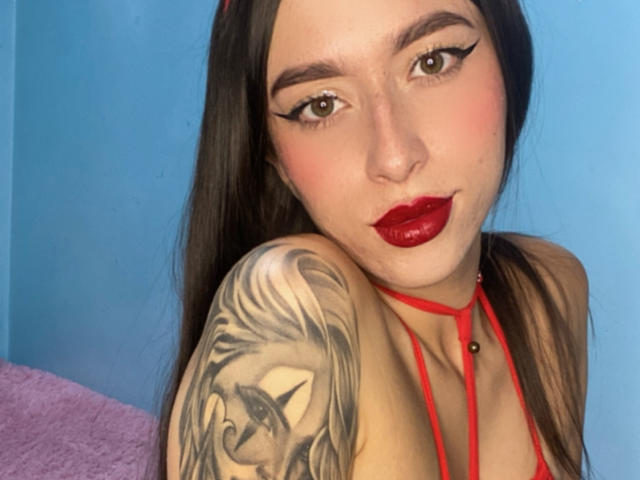 SarahLuxe with red lipstick, black eyeliner, and a tattoo of a unicorn on her right shoulder, wearing a red strappy top, against a blue wall.
