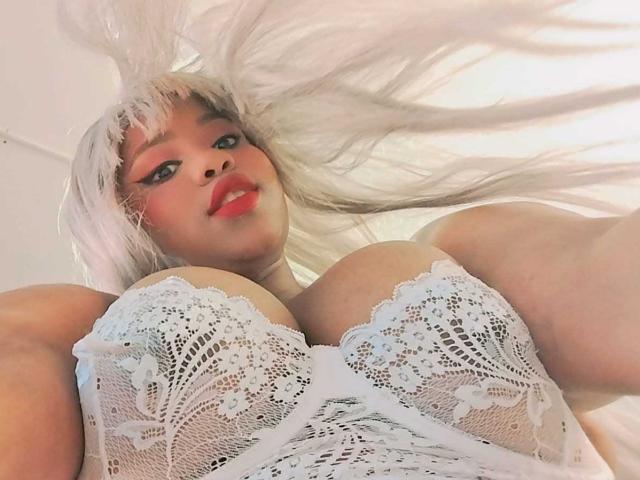 LizzyFoxy with huge tits in a white lace bra, looking down at the camera with long white hair floating. She's smiling with red lipstick, and her eyes are looking up.