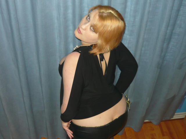 SeductiveBodyX with short blonde hair, black top, and black pants, showing her bare ass while looking over her shoulder, in a room with blue curtains.