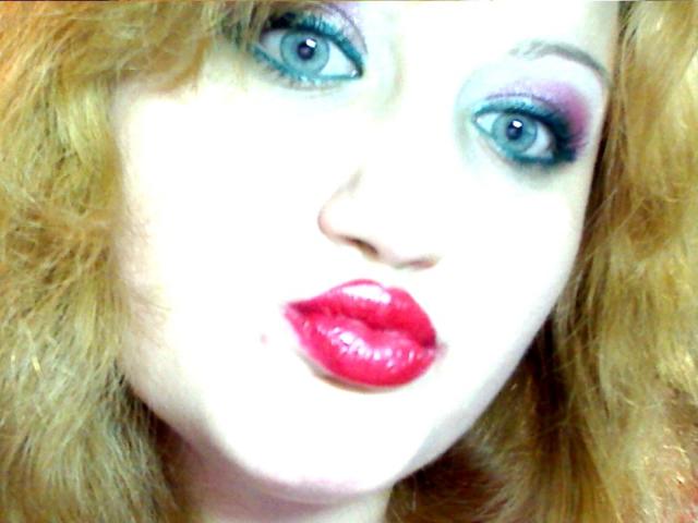SeductiveBodyX has blue eyes, heavy purple and black eyeshadow, red glossy lips, and wavy blonde hair. Her face is slightly pale with a small zit near her mouth.