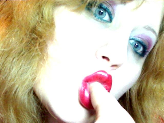 SeductiveBodyX with bright blue eyes, pink glossy lips, and red lipstick, holding a pink candy near her mouth. She has wavy blonde hair and white makeup around her eyes.