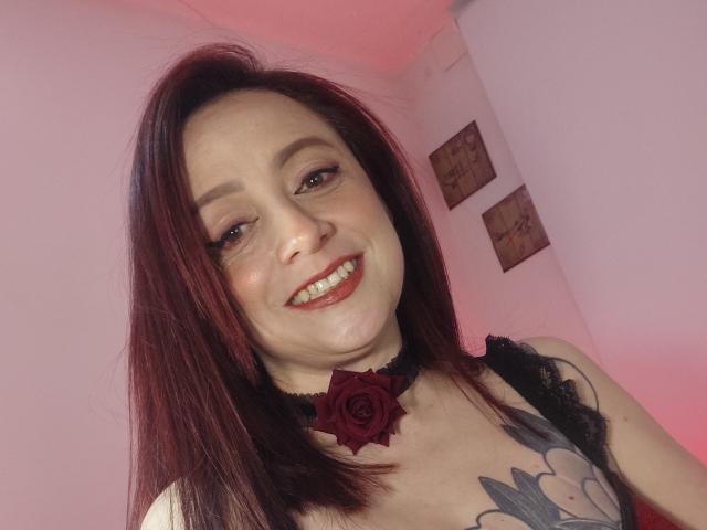 CatalinaMonyque with red hair, smiling, wearing a black choker with a rose, showing tattoos on her chest, and white top, looking at the camera.