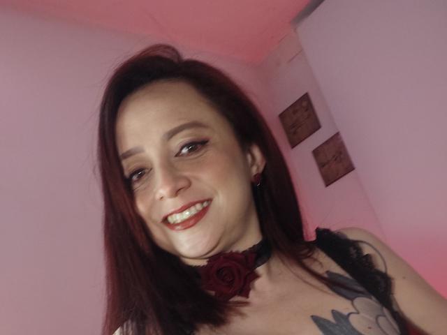 CatalinaMonyque with red hair, smiling, wearing a black choker with a red rose, tattoo on chest, dark eyeliner, and red lipstick, in a pink room.
