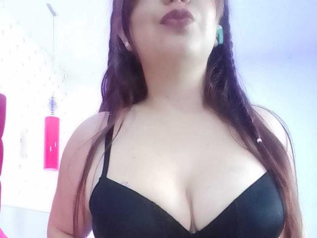 AmatisthaBlend with pale skin, dark brown hair, and large boobs in a black bra, wearing green earrings, has full lips with dark lipstick. Background is blurred.
