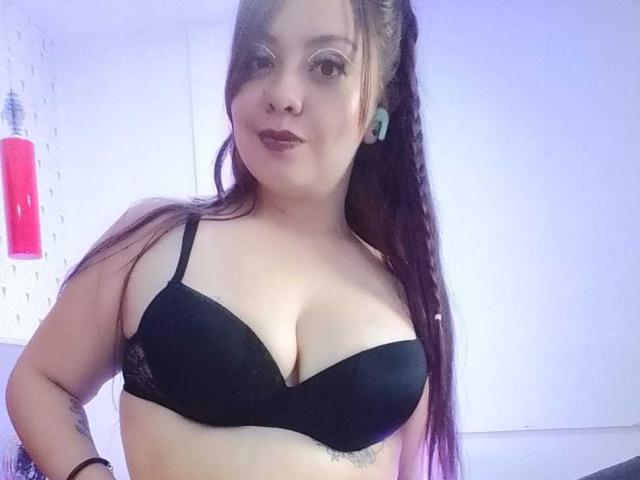 AmatisthaBlend with long brown hair, wearing a black bra, showing big tits, light skin, small tattoo below her chest, blue earbuds, and dark lipstick, in a dimly lit room.